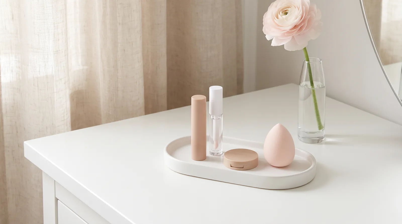 Penteadeira branca com bandeja de poucos itens de maquiagem (stick, gloss, estojo compacto e esponja) ao lado de uma flor rosa