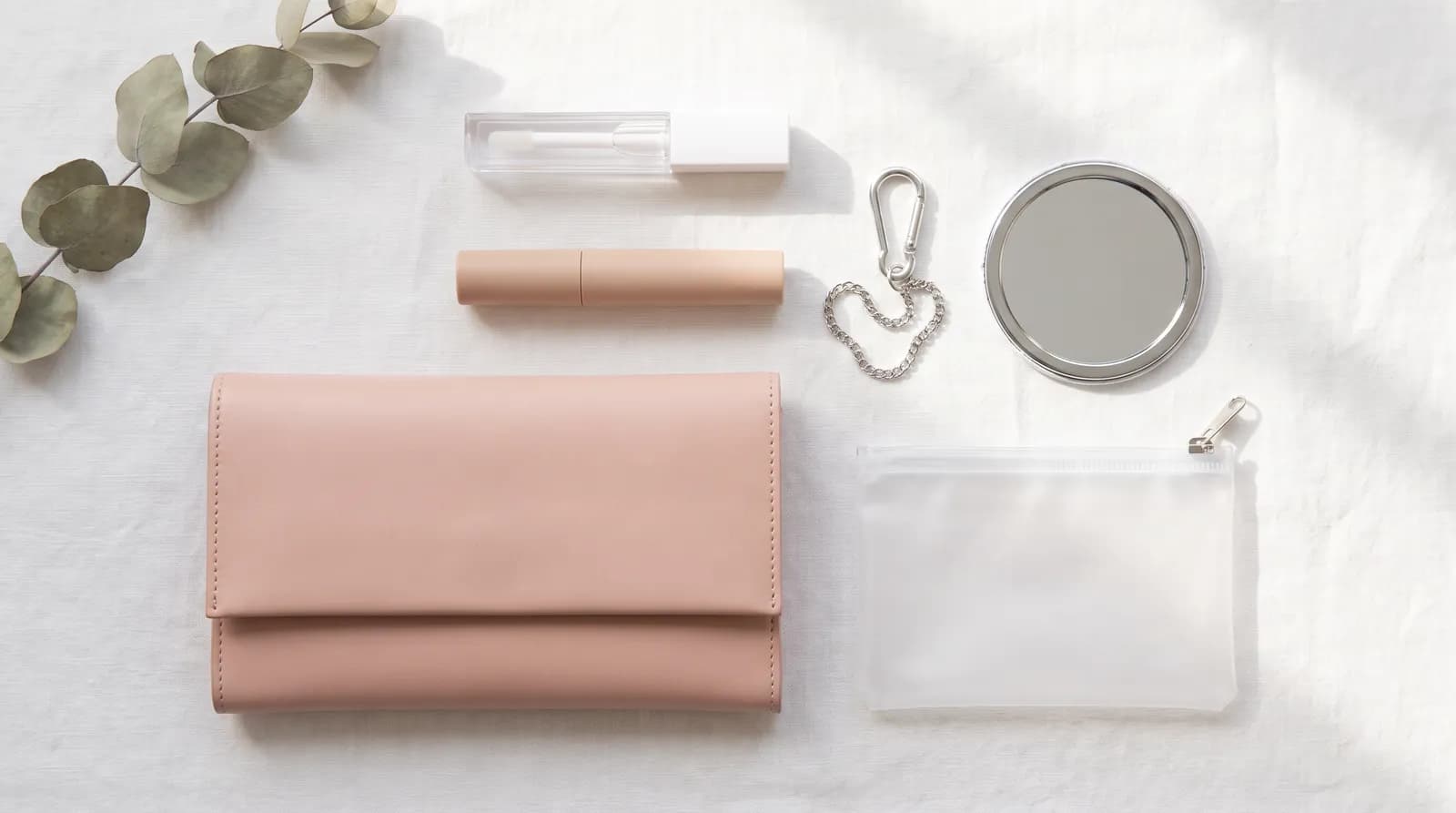 Itens para levar na bolsa sobre tecido branco: carteira rosa, nécessaire fosca, espelho de bolso, gloss, stick de maquiagem e mosquetão com corrente